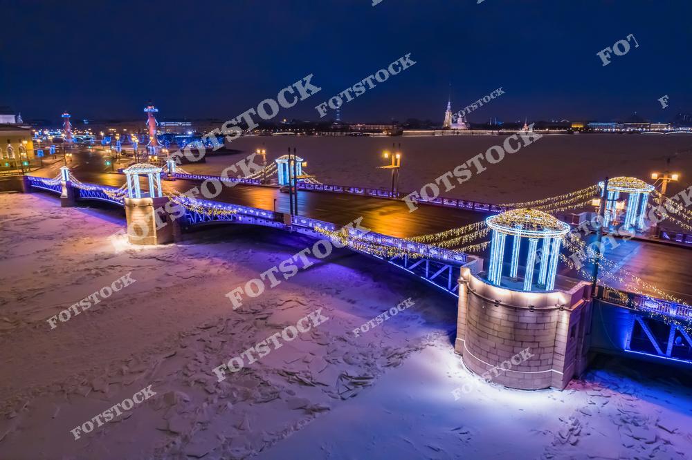 На фотографии изображён Дворцовый мост — один из самых знаменитых мостов Санкт-Петербурга, украшенный новогодней иллюминацией. На левом берегу виднеются Ростральные колонны и здание Биржи на Стрелке Васильевского острова. Справа за Невой — Петропавловская крепость с собором святых Петра и Павла. Лёд на реке подчёркивает суровую и красивую зиму Северной столицы. Праздничные огни создают атмосферу уюта и торжества.